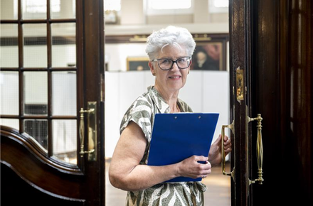 A smiling woman with short white hair and glasses holds a blue clipboard while standing at the entrance of a formal hall.